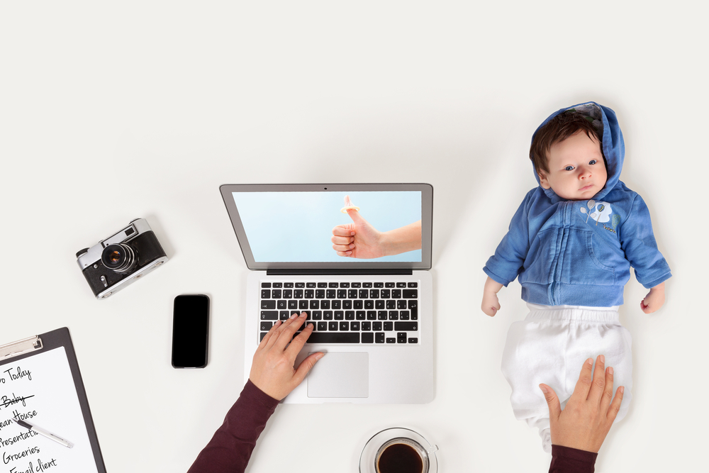Mother holding baby while working on laptop in office. Business and family balance. Workplace with baby, laptop, phone and to do list from above.